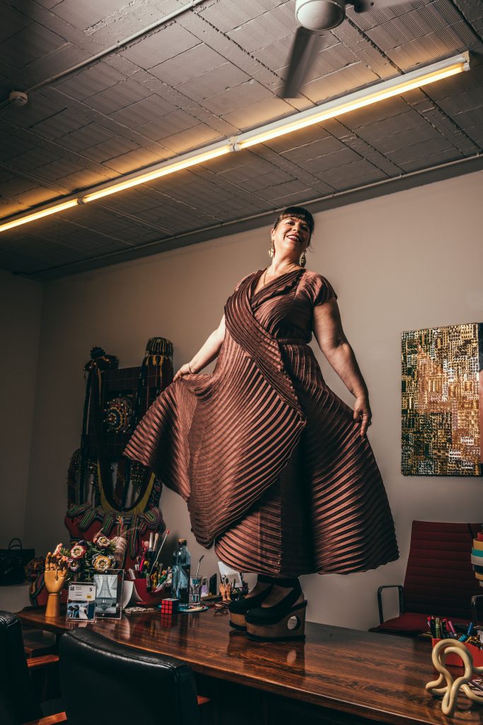 Portrait of Joana Vasconcelos standing ona. table in a brown dress surrounded by various artmaking objects.